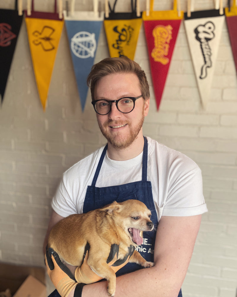 Man holding a small dog wearing an apron with pennants in the background
