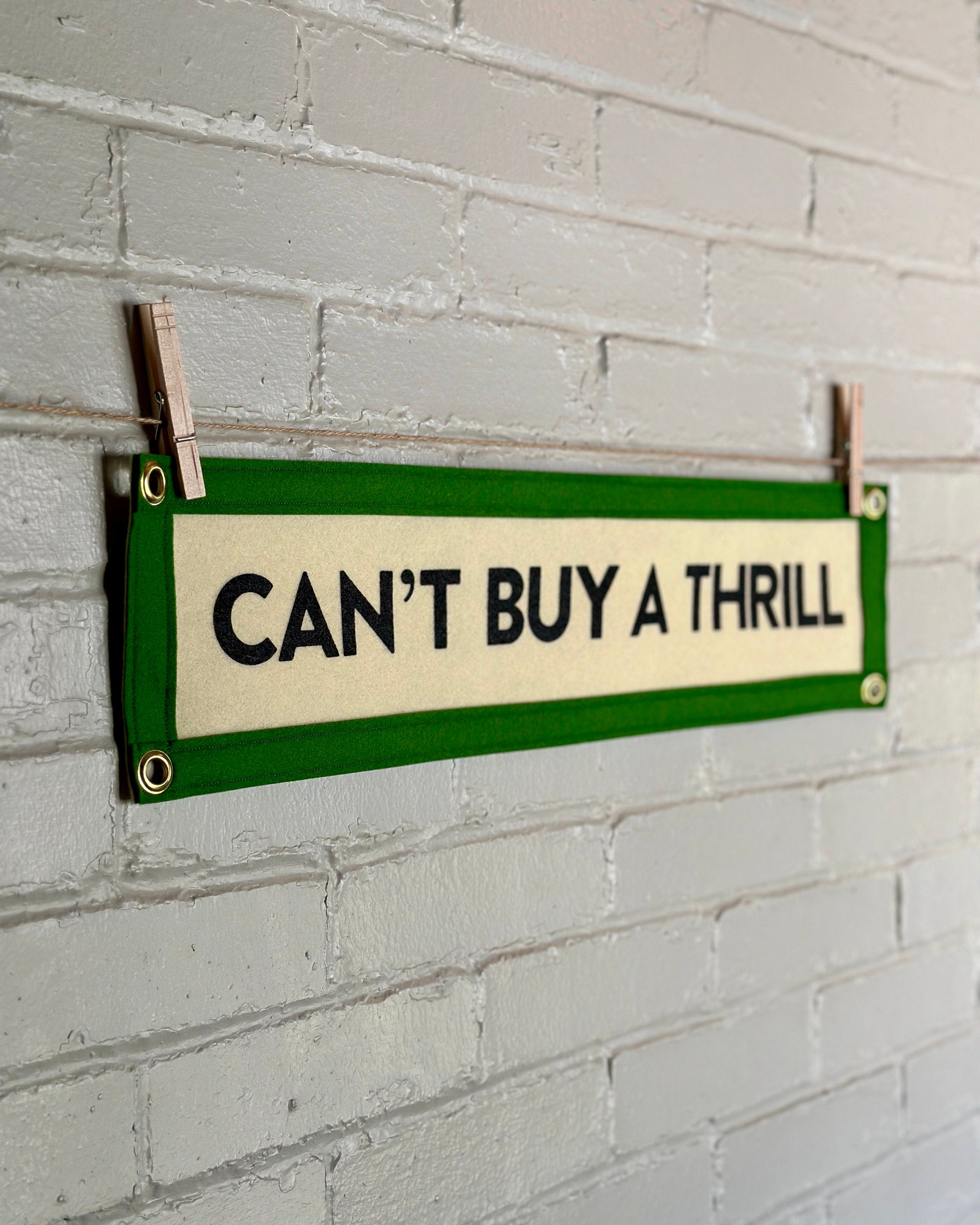 Side view of a cream felt banner with green trim and black lettering that reads "Can't Buy a Thrill"