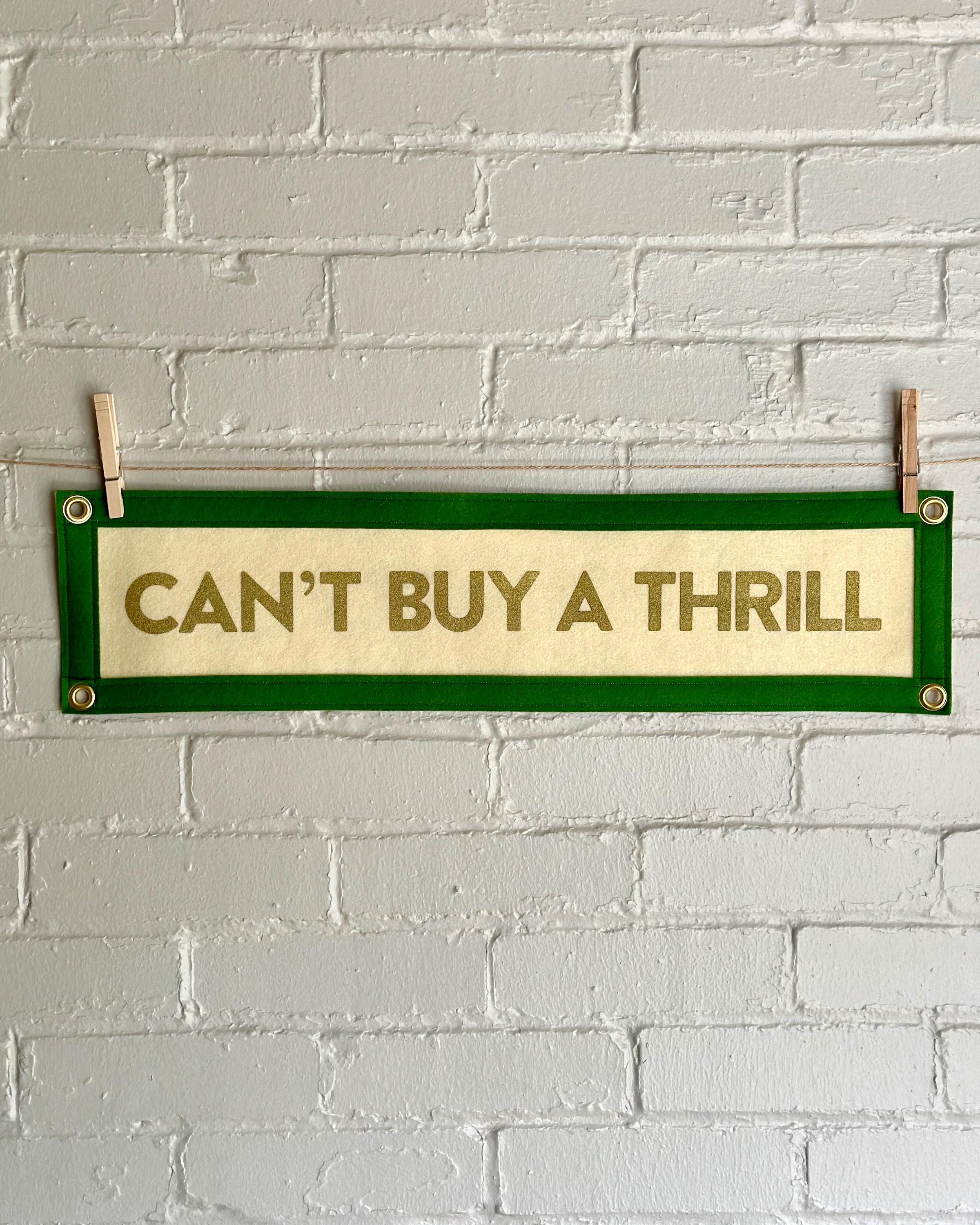 A cream felt banner with green trim and gold lettering that reads "Can't Buy a Thrill"