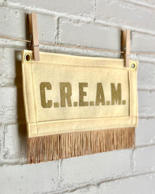 Side view of a cream felt banner with cream trim and gold fringe, and gold lettering that reads 'C.R.E.A.M.'