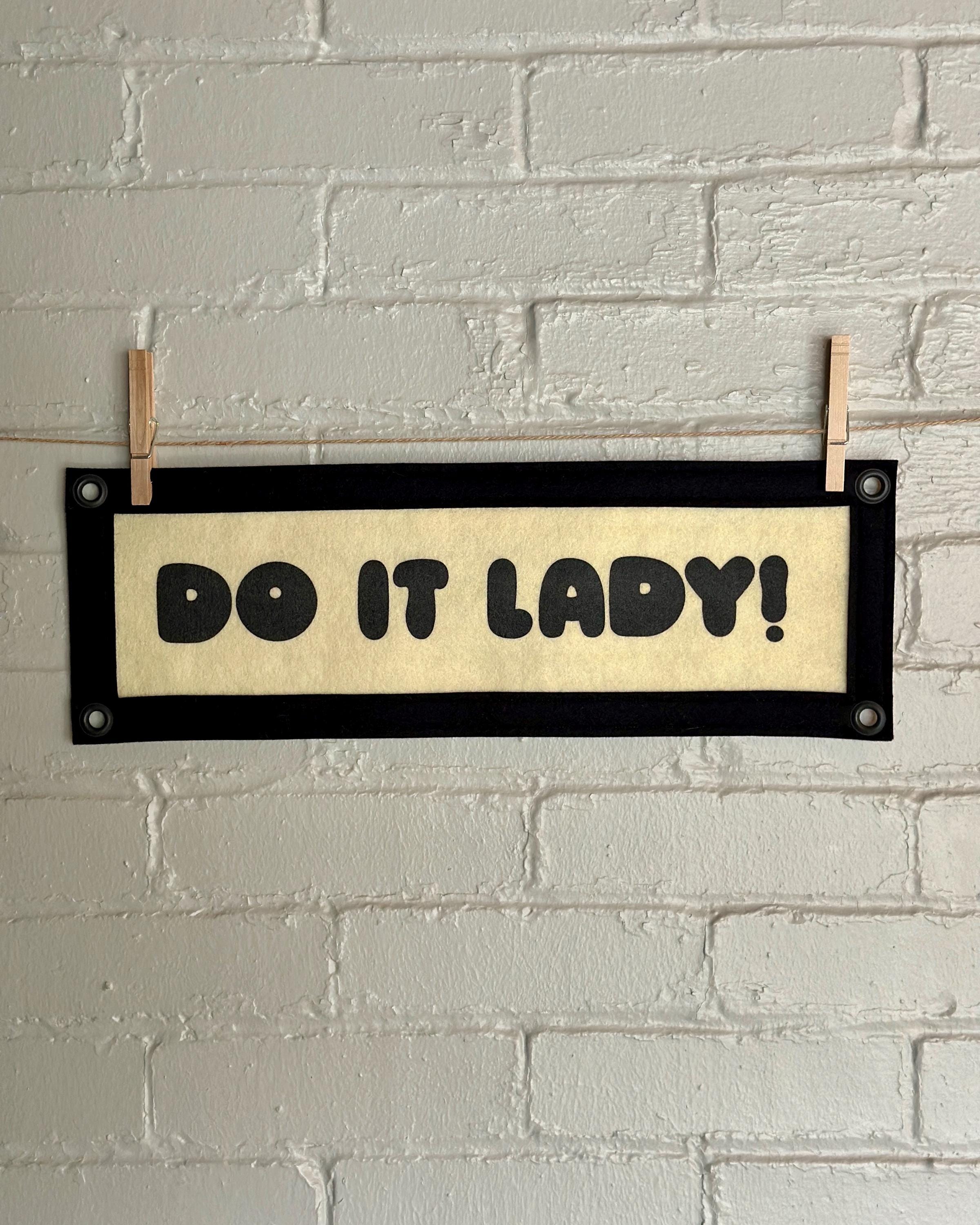 A cream felt banner with black trim and lettering that reads "Do it lady!"