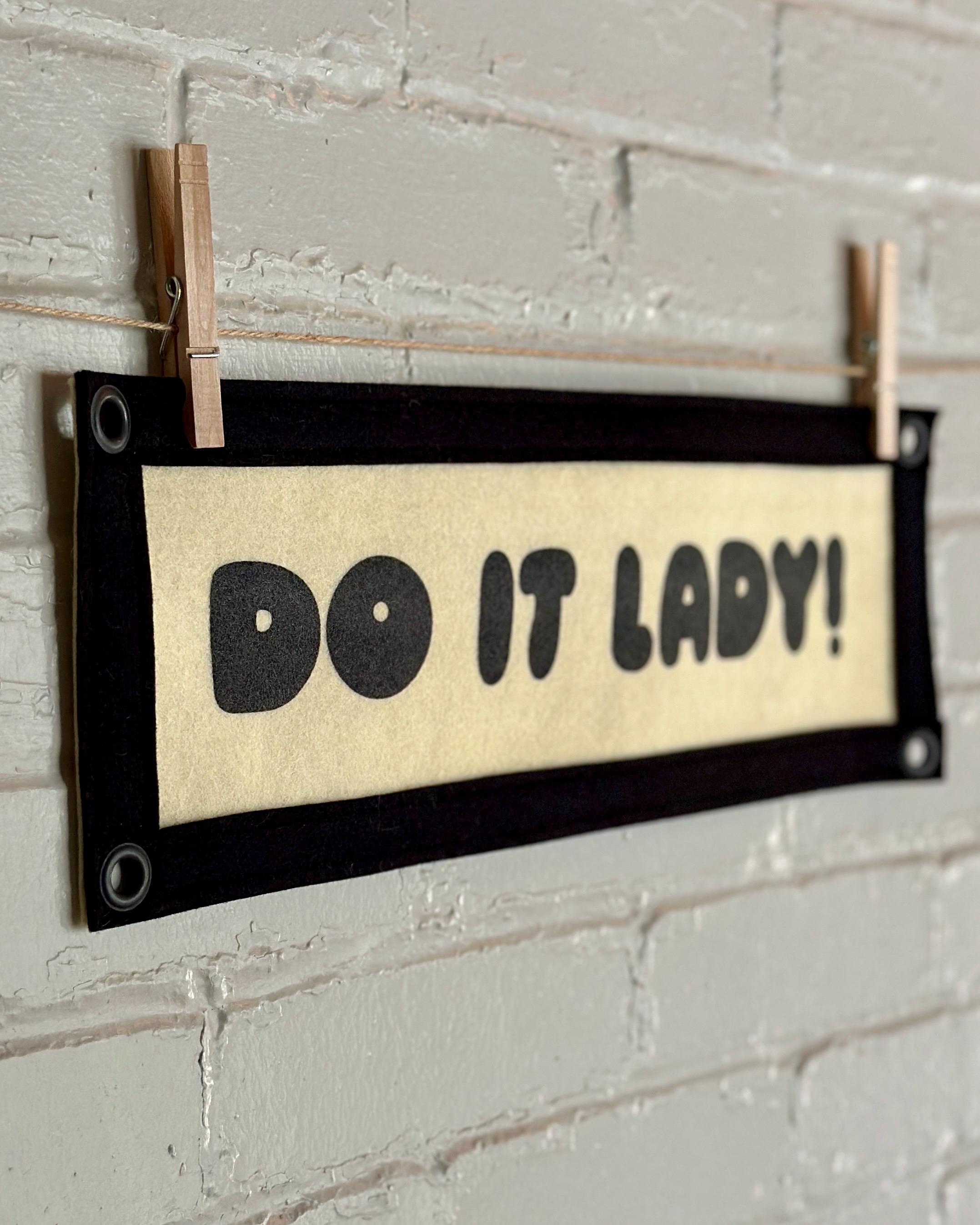 Side view of a cream felt banner with black trim and lettering that reads "Do it lady!"