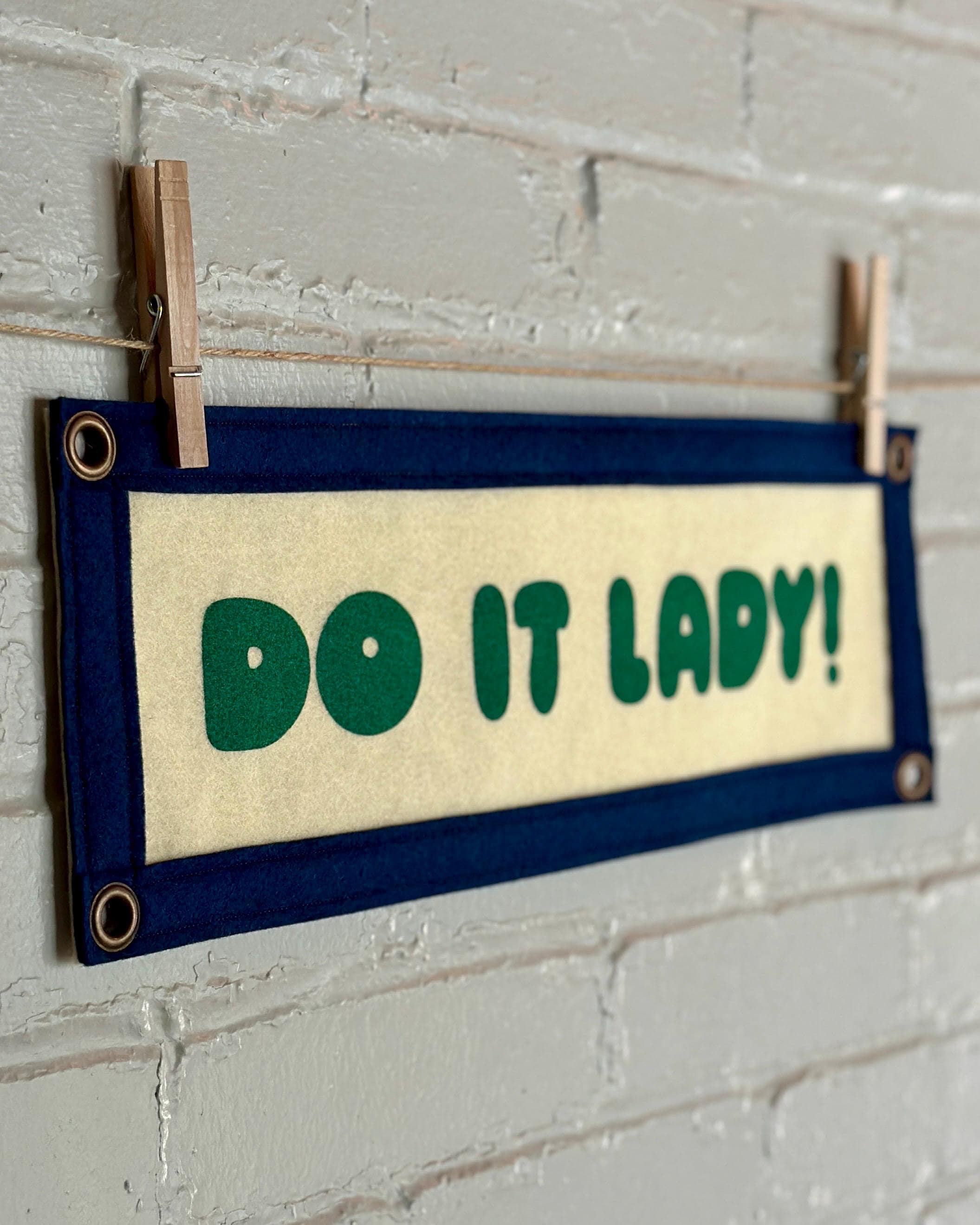 Side view of a cream felt banner with navy trim and green lettering that reads "Do it lady!"