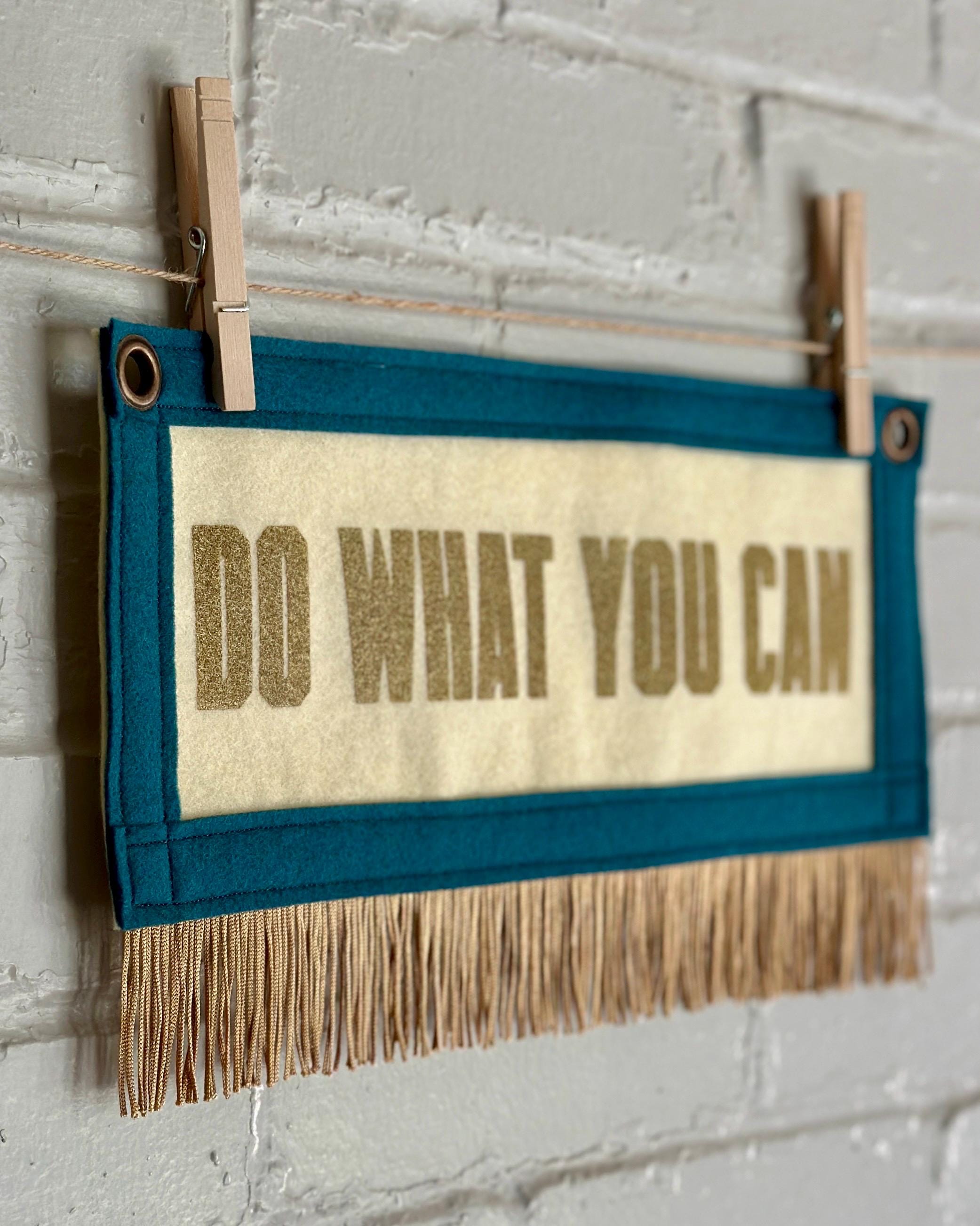 Side view of a cream felt banner with blue spruce trim, gold fringe and lettering that reads "Do what you can"