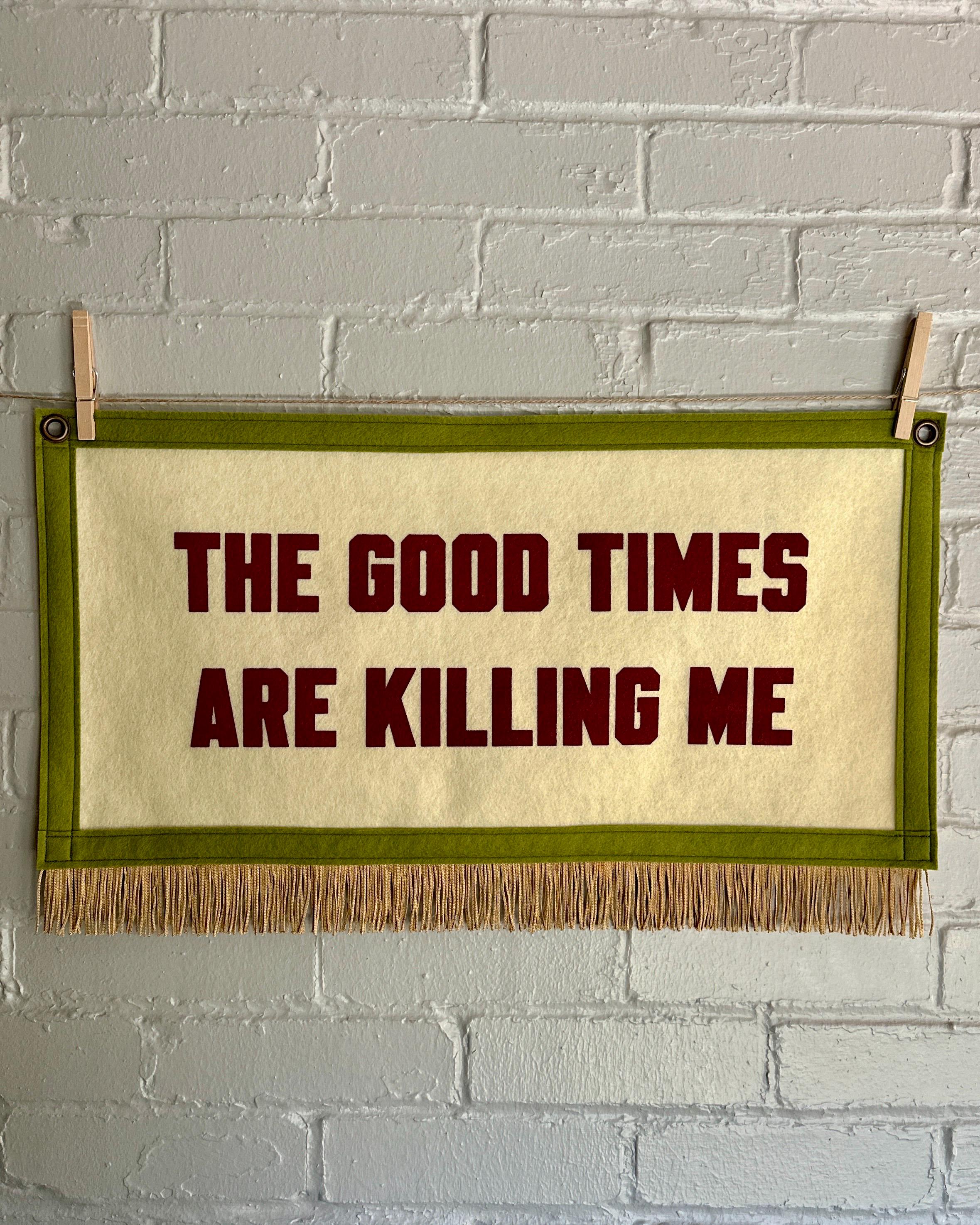 A large cream banner with pea green felt trim, gold fringe, and blood red lettering that reads "The good times are killing me"