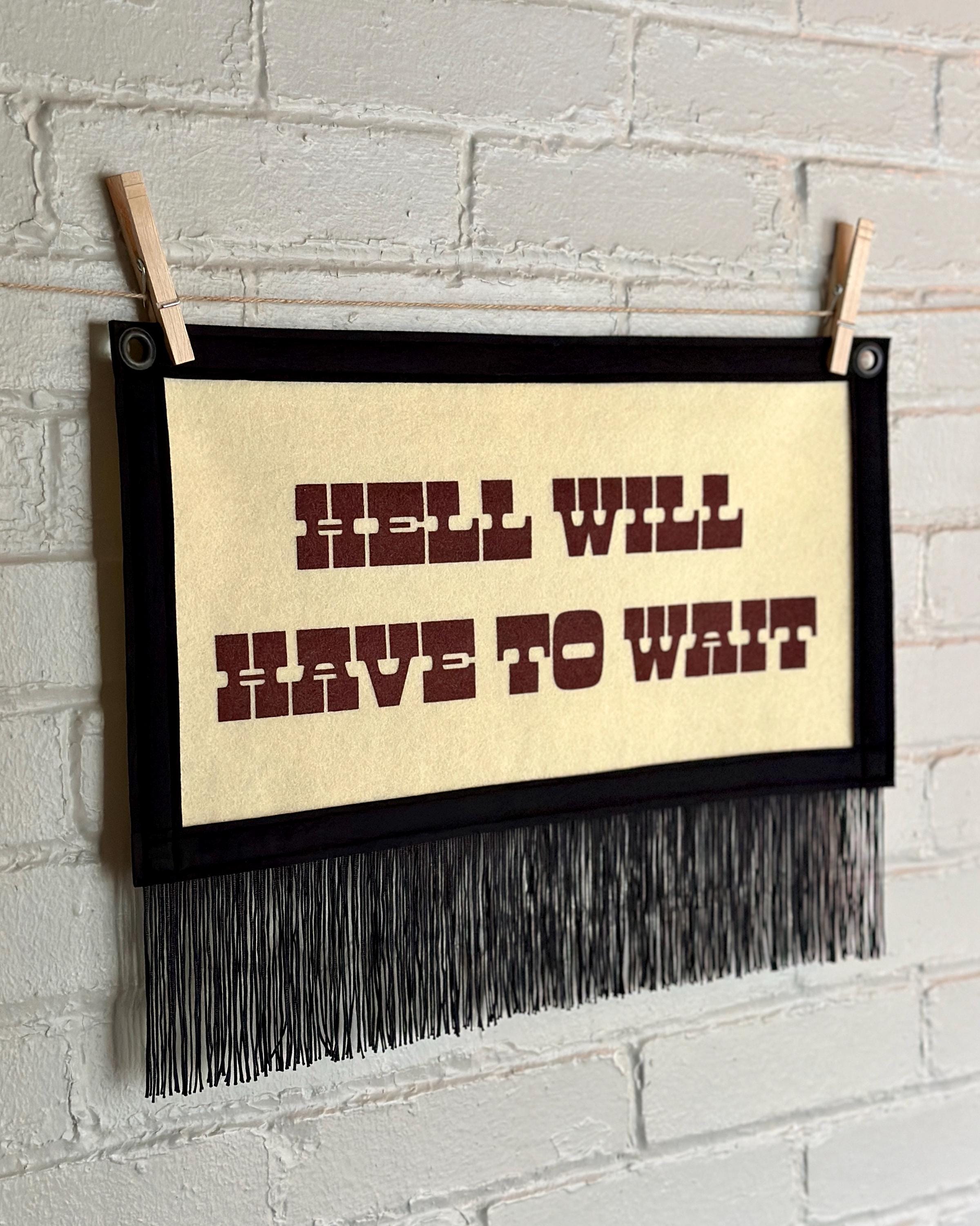 Side view of a cream felt banner with black trim, long black fringe, and blood red lettering reading "Hell will have to wait"