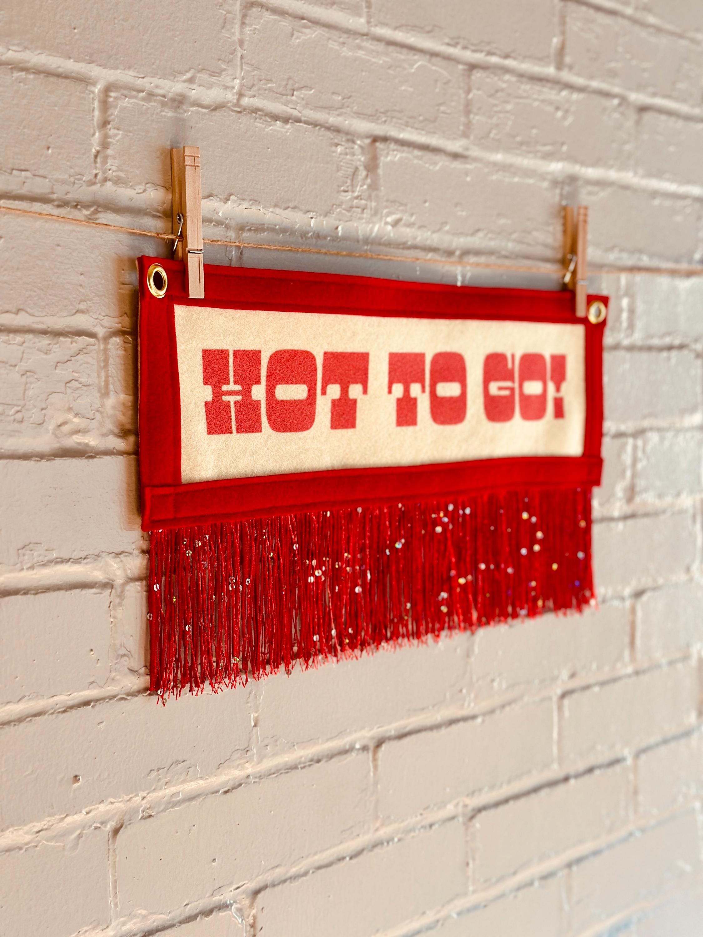 Side view of a cream felt banner with red trim and red sequin fringe, pink lettering reads "HOT TO GO!"