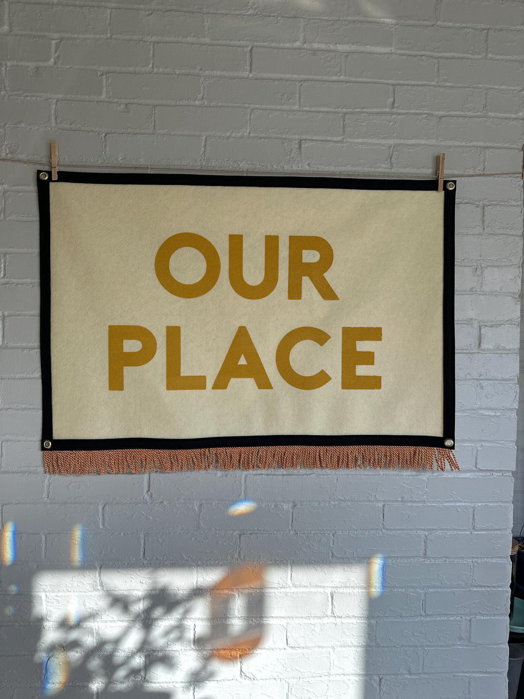 A large cream felt banner with black trim, gold bullion fringe, and letters in yellow ink that read "OUR PLACE"