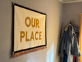 A dining room wall with coat rack and large cream felt banner with black trim, gold bullion fringe, and letters in yellow ink that read "OUR PLACE"