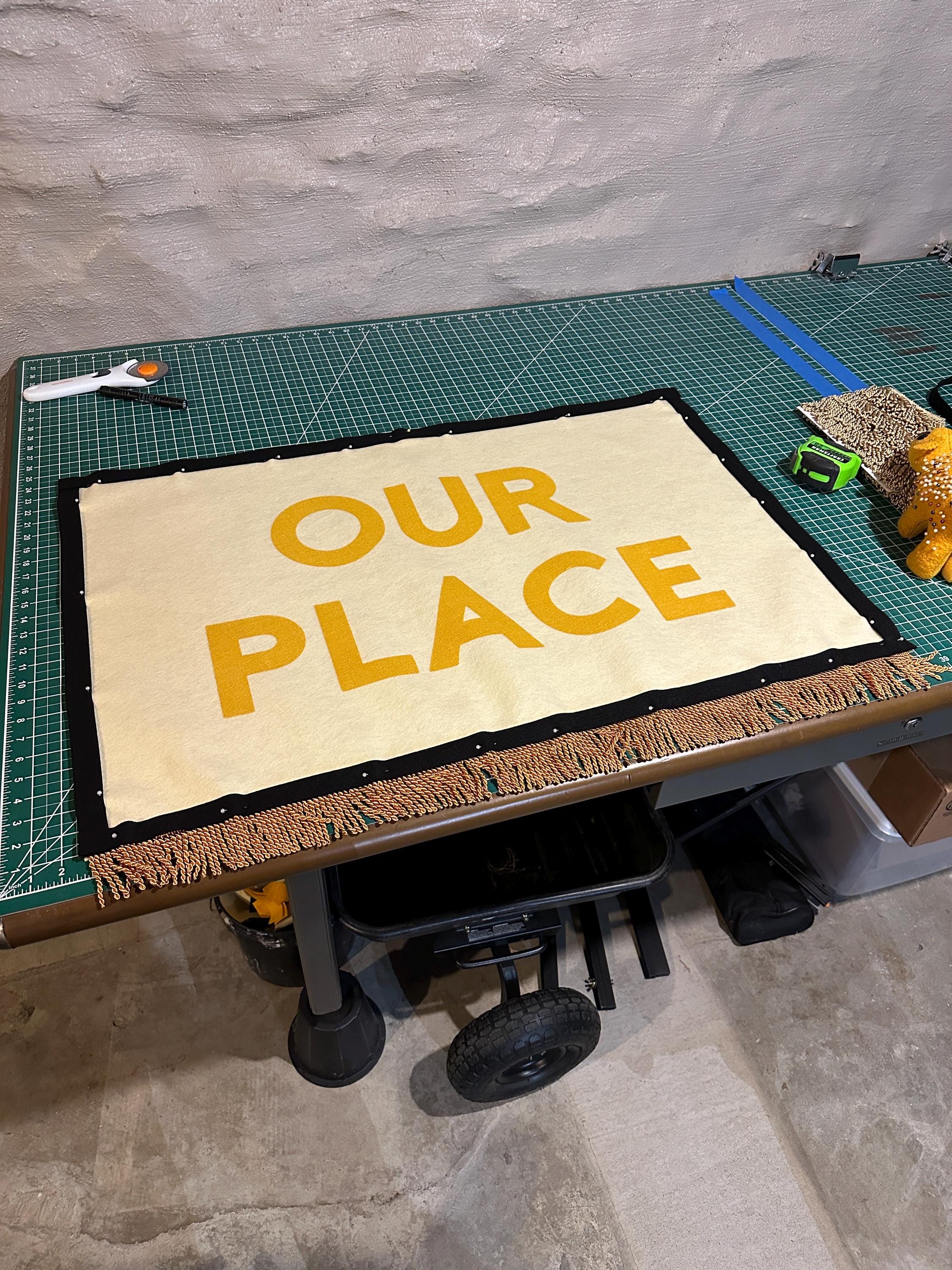 An OUR PLACE banner in progress on a large table and cutting mat with trim attached with sewing pins