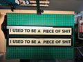 Two cream felt banners with black trim lettering reading "I USED TO BE A PIECE OF SHIT" sitting on top of a cutting mat. A T square is depicted showing a slightly crooked misaligned print