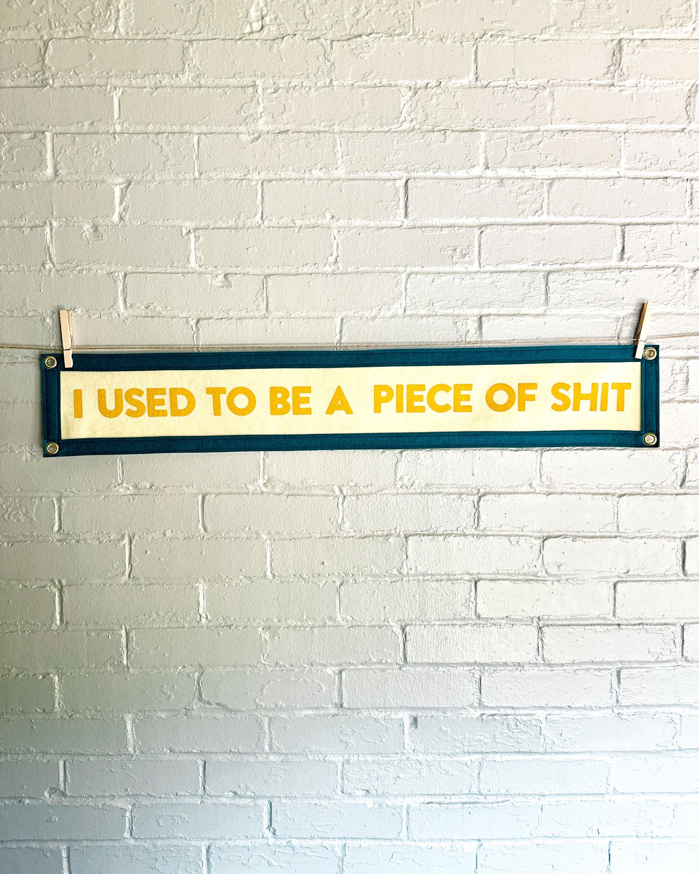 A cream banner with spruce blue trim and yellow lettering that reads "I used to be a piece of shit"