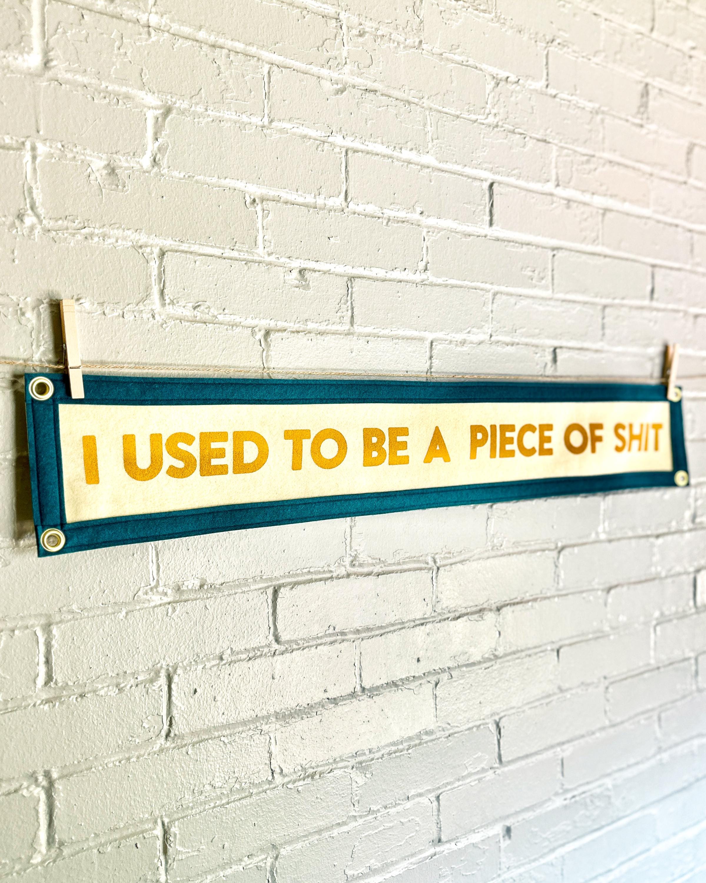 Side view of a cream banner with spruce blue trim and yellow lettering that reads "I used to be a piece of shit"