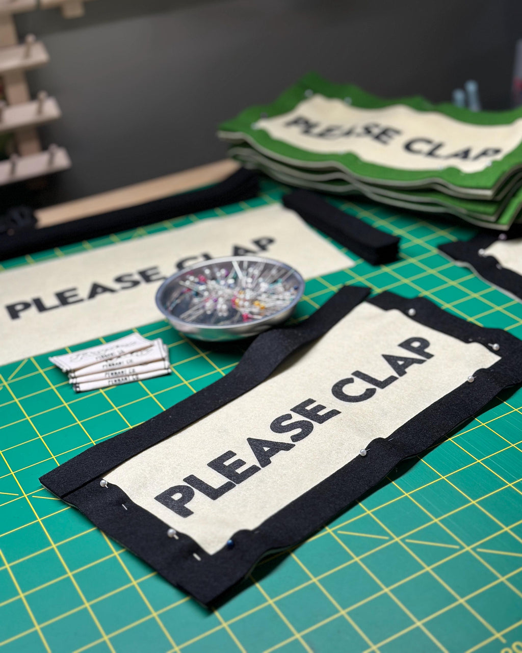 Various felt banners and components on a desk cutting mat beside sewing pins and Payday Pennant Co. tags
