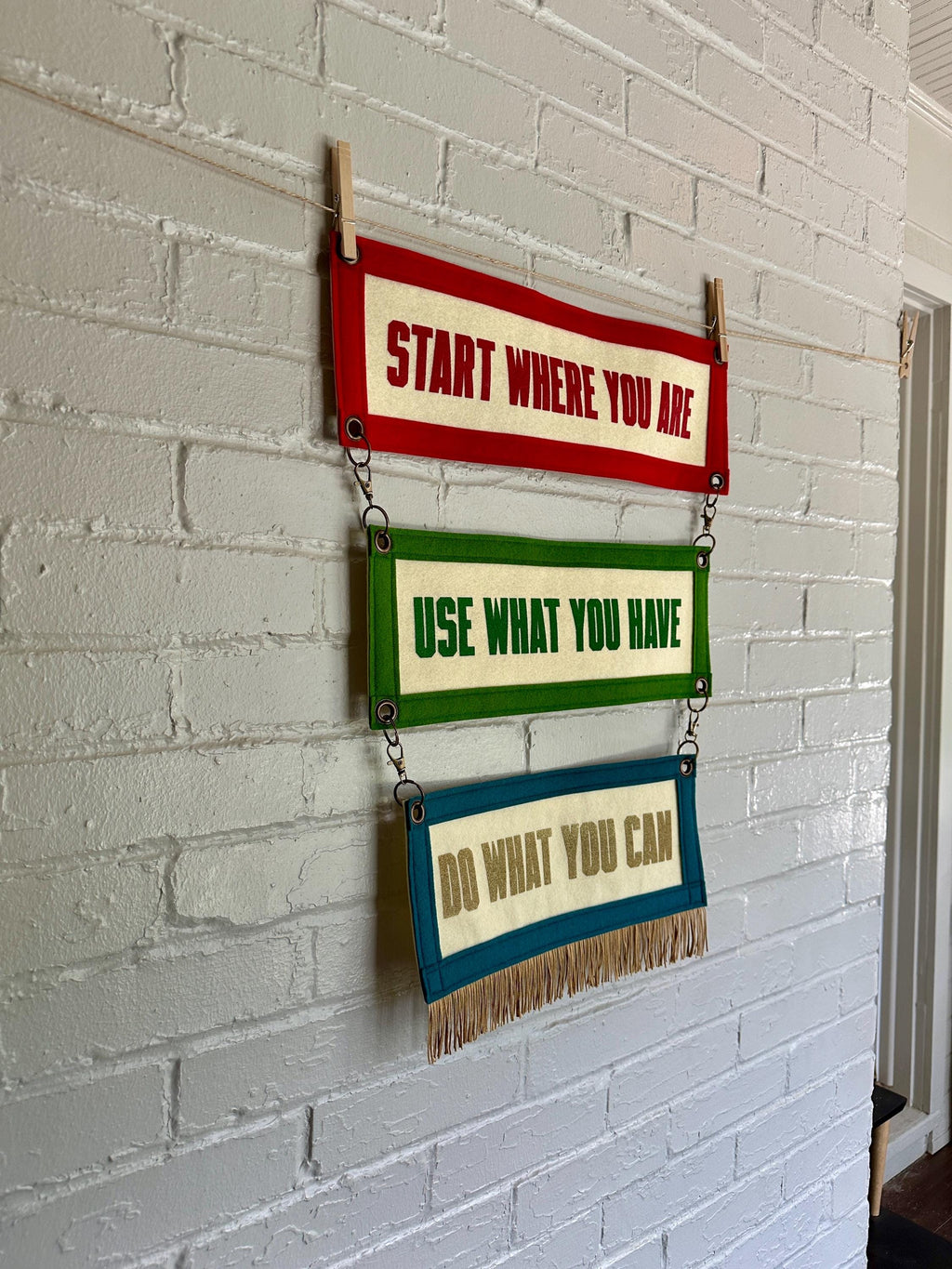 Side view of three colorful felt banners with motivational phrases attached by brass rings, hanging on a white brick wall
