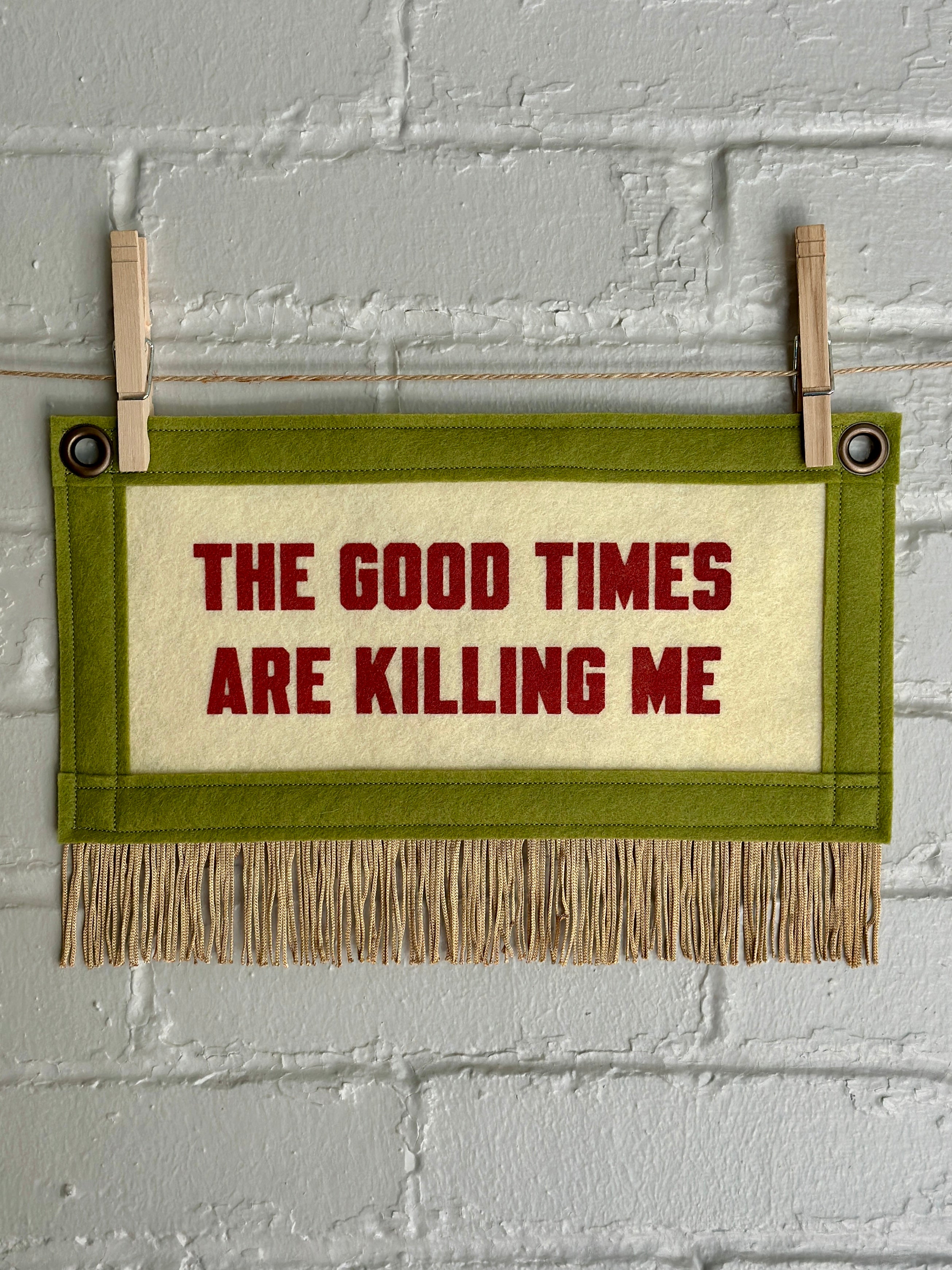 A small cream felt banner with green trim and gold fringe at the bottom. Words read "The good times are killing me" in red ink.