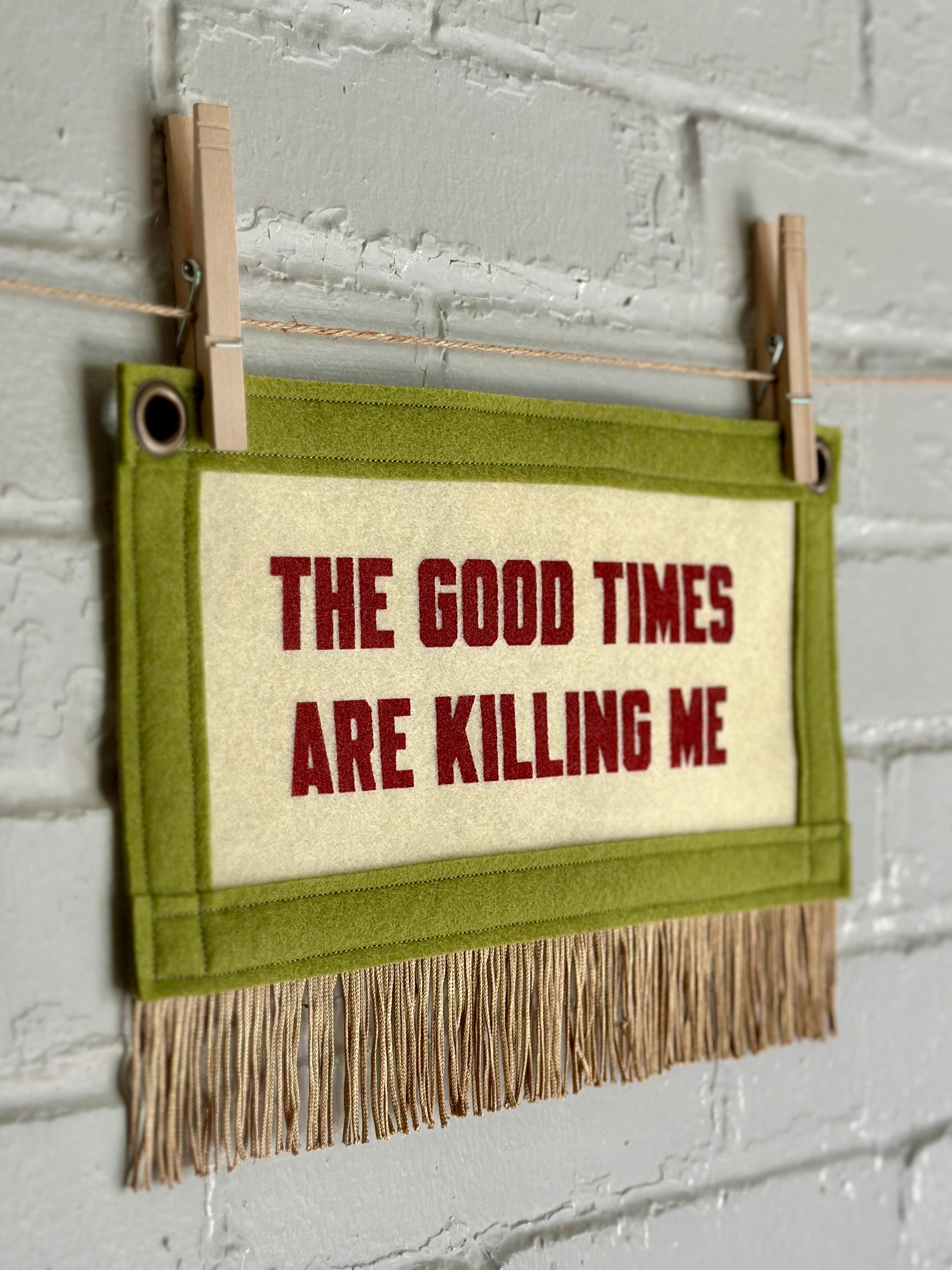 A small cream felt banner with green trim and gold fringe at the bottom. Words read "The good times are killing me" in red ink.