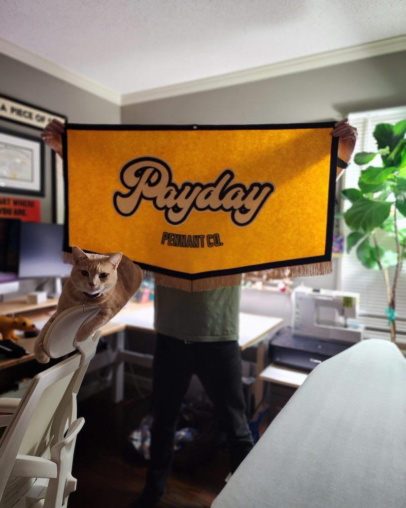 Person holding a large yellow 'Payday Pennant Co.' banner with a cat sitting on a chair in a room.