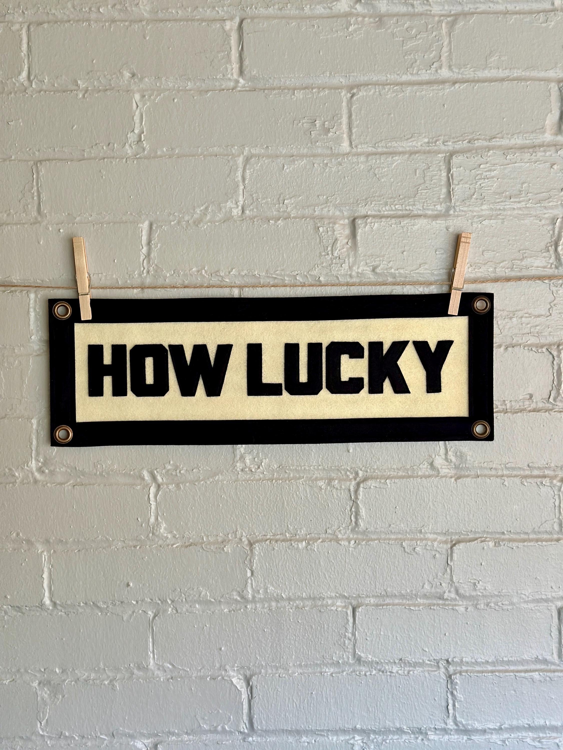 A cream felt banner with black stitched letters that read 'HOW LUCKY' hanging from two wooden clothespins against a white brick wall.