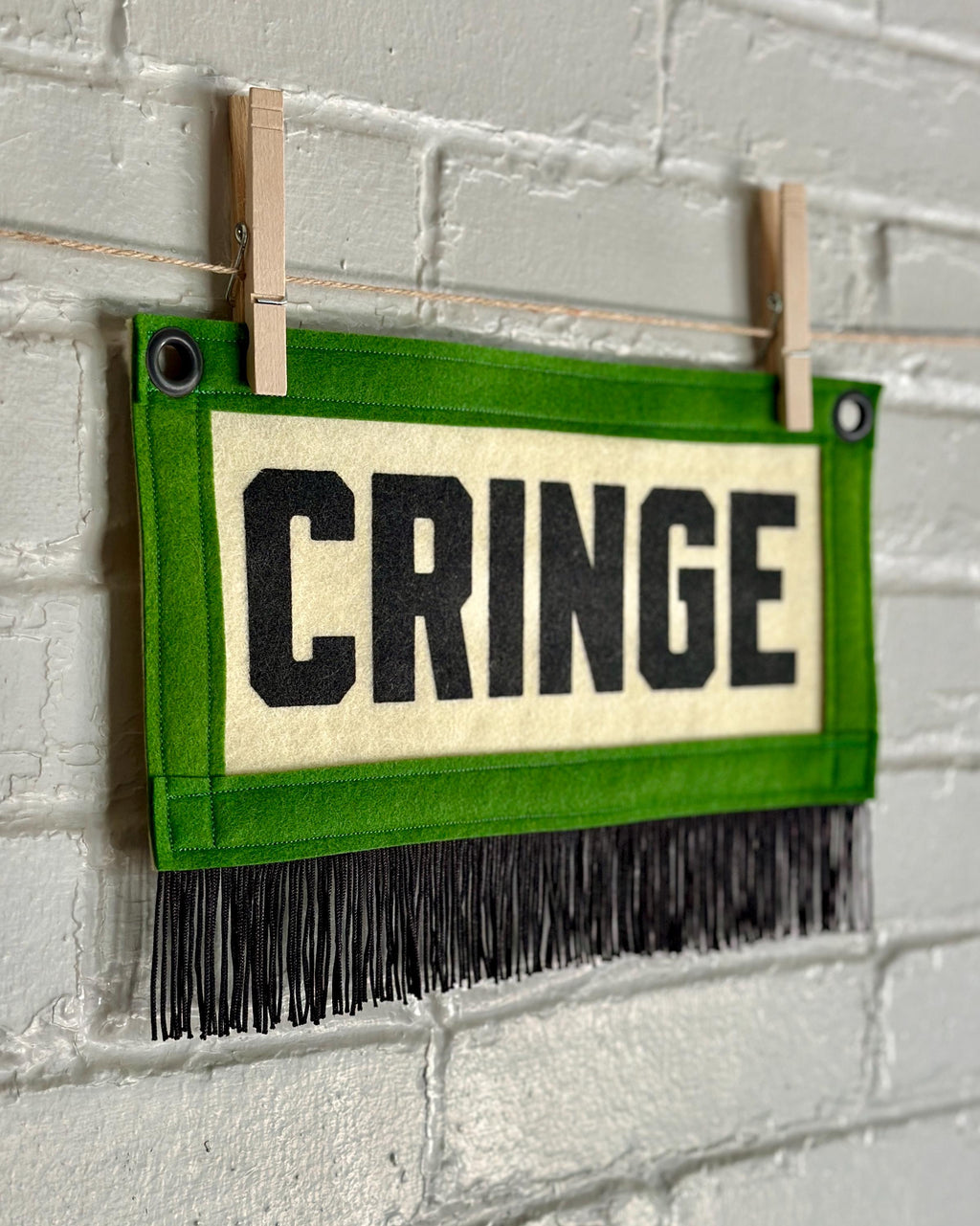 A side view of a small felt banner with cream base, green trim and black fringe at the bottom. It reads CRINGE in large letters with black ink.