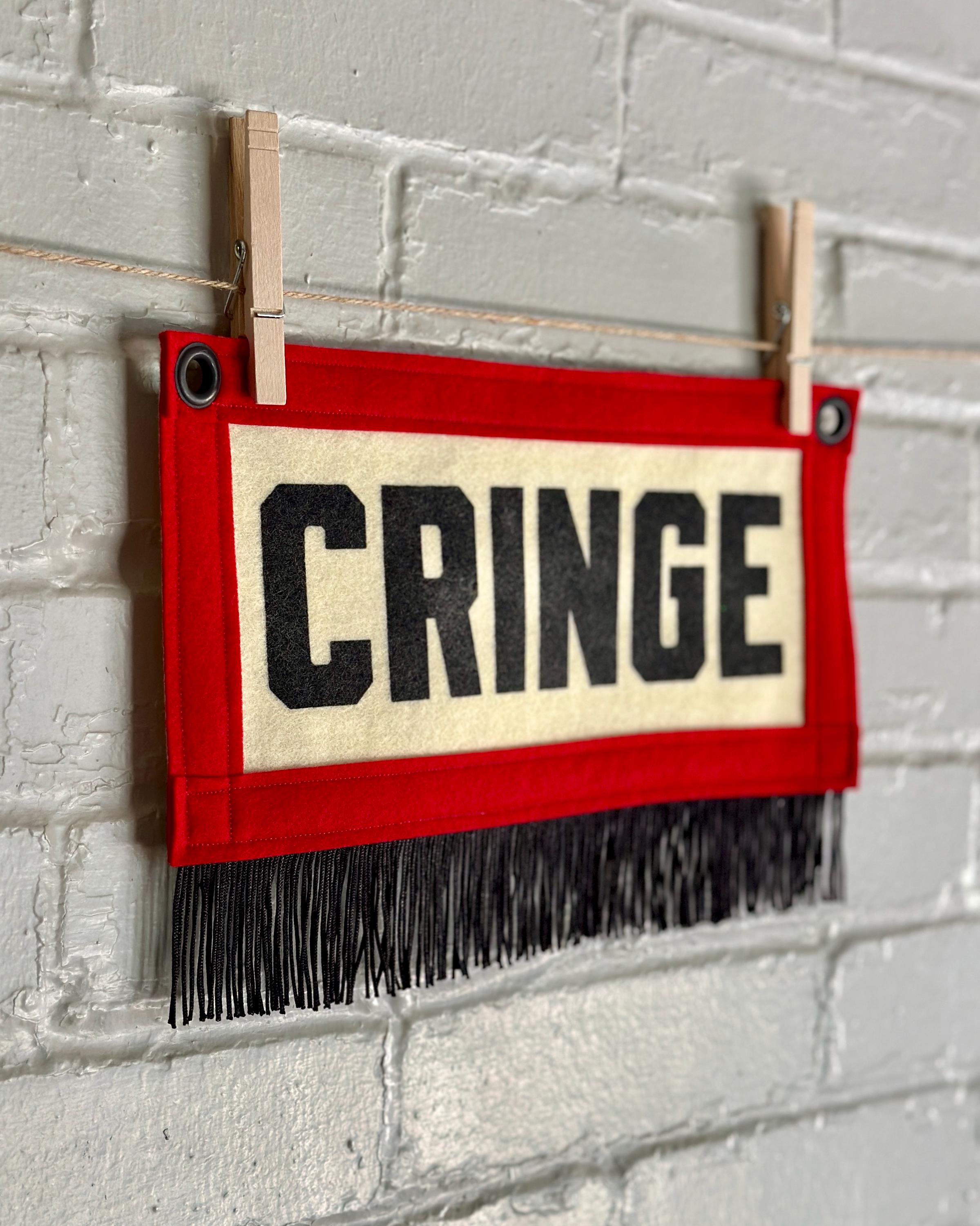 A side view of a small felt banner with cream base, red trim and black fringe at the bottom. It reads CRINGE in large letters with black ink.