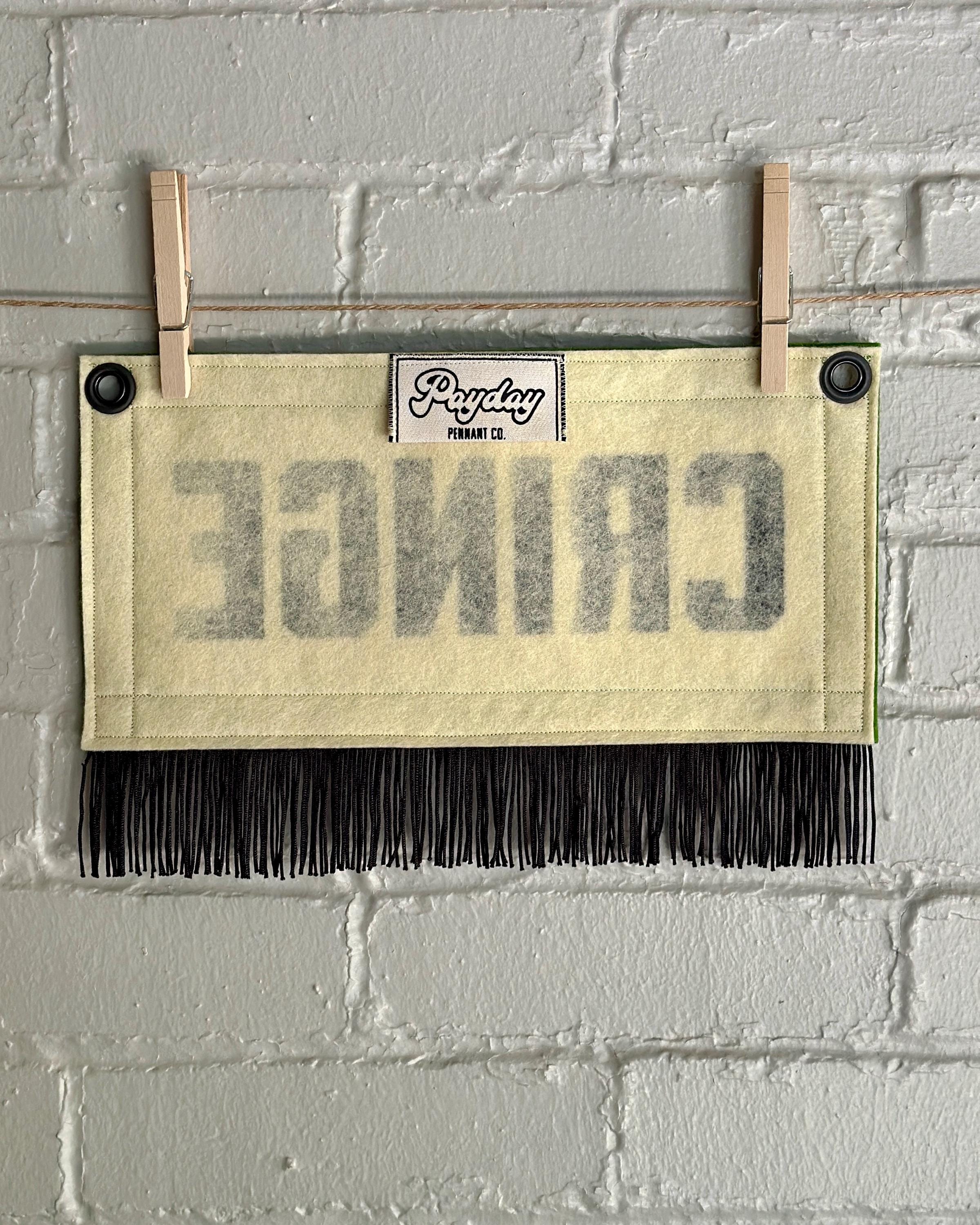 The back of a small felt banner with cream base, green trim and black fringe at the bottom. The back of the banner shows a Payday Pennant Co. tag.