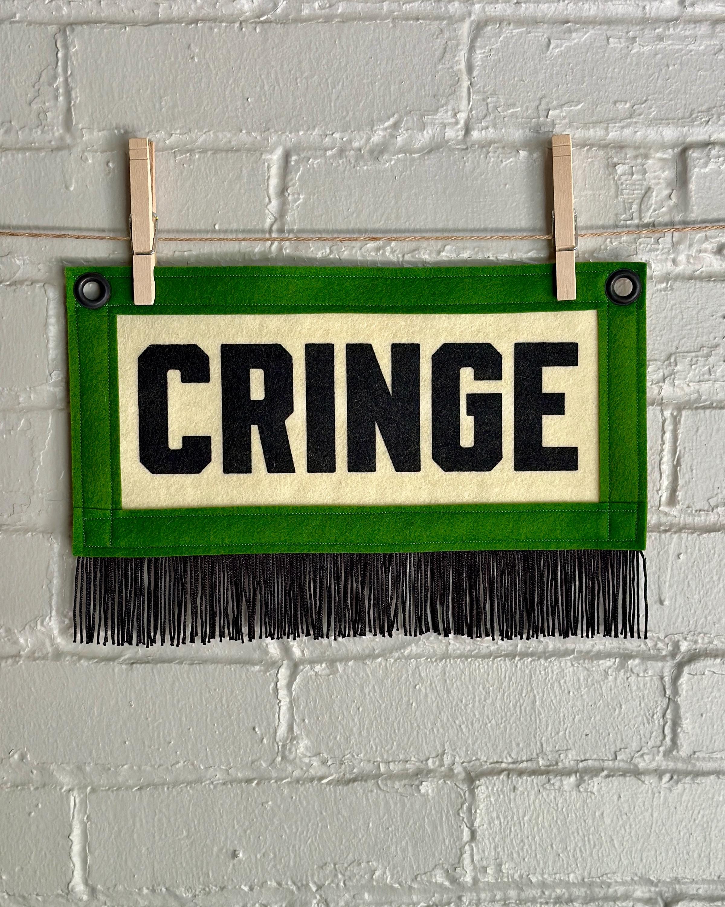 A small felt banner with cream base, green trim and black fringe at the bottom. It reads CRINGE in large letters with black ink.