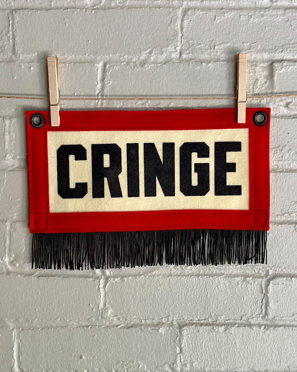 A small felt banner with cream base, red trim and black fringe at the bottom. It reads CRINGE in large letters with black ink.