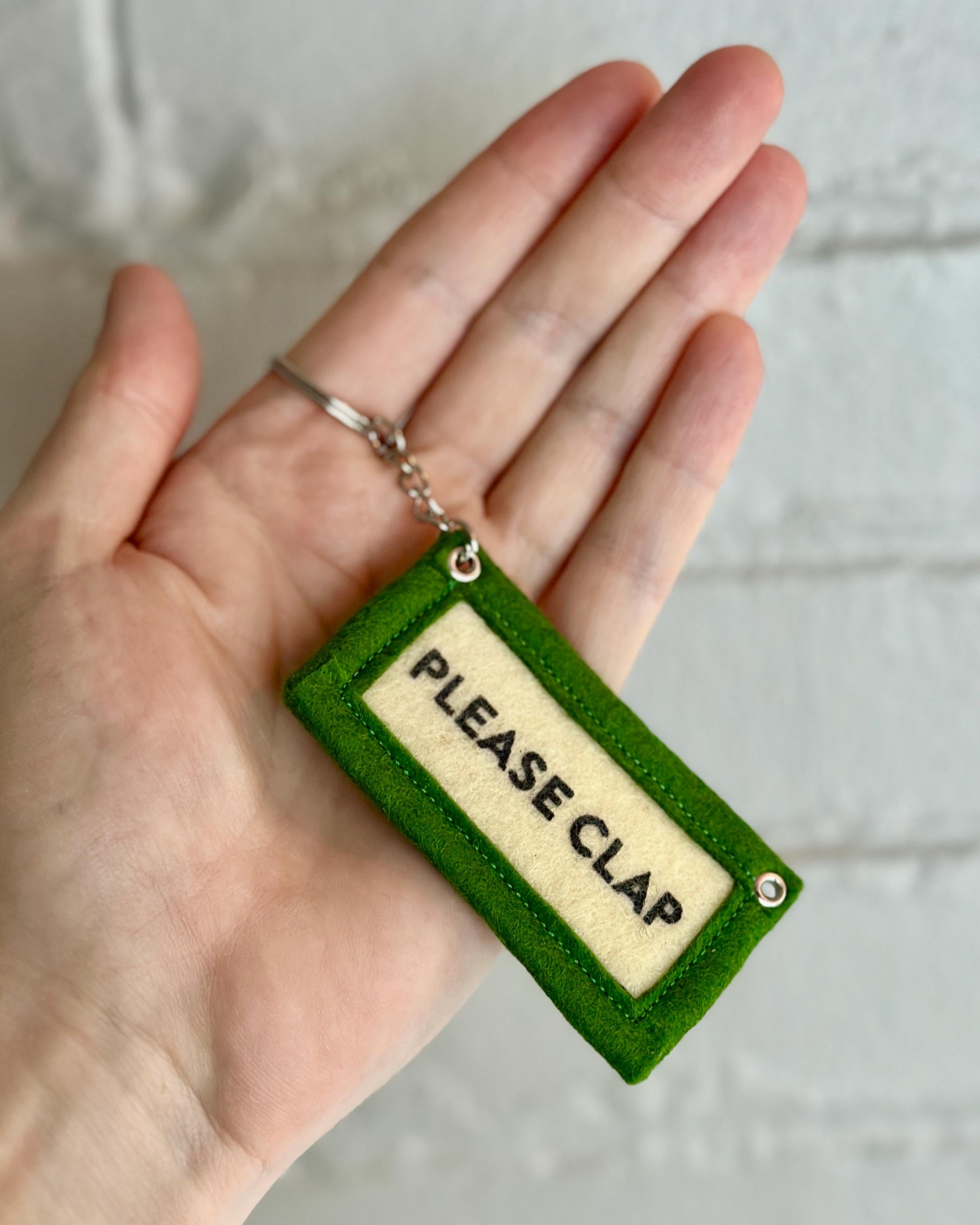 Hand holding a green felt keychain with 'PLEASE CLAP' text