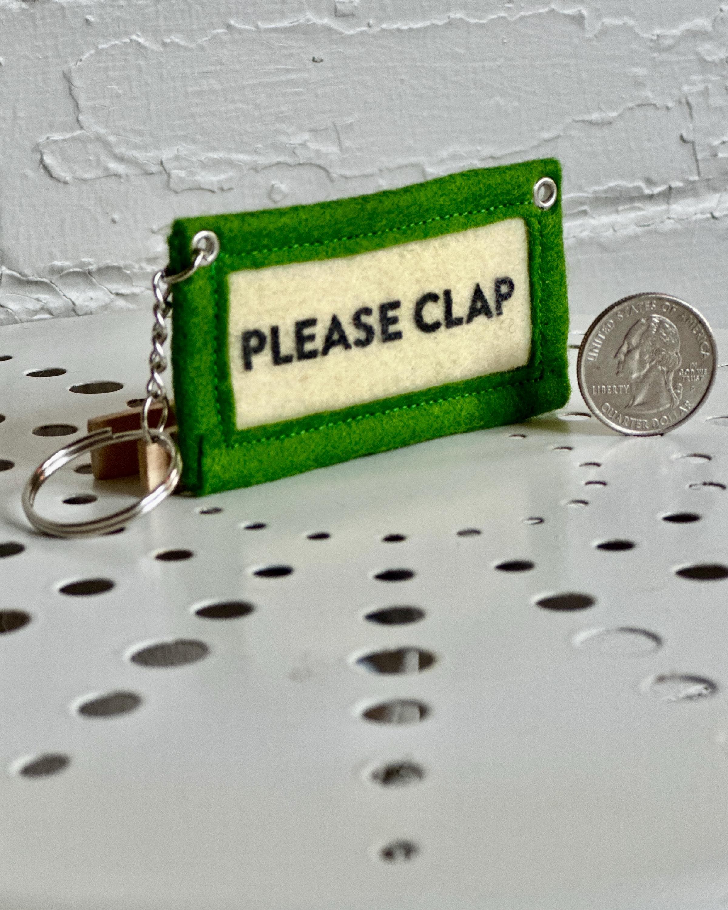 Green felt keychain with 'PLEASE CLAP' text sitting next to a US quarter for scale