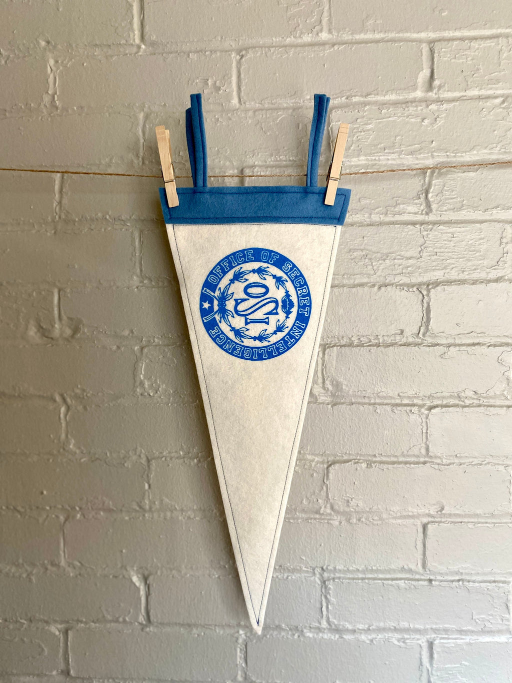 A white felt pennant with light blue trim and the OSI logo from the cartoon The Venture Bros printed in blue ink