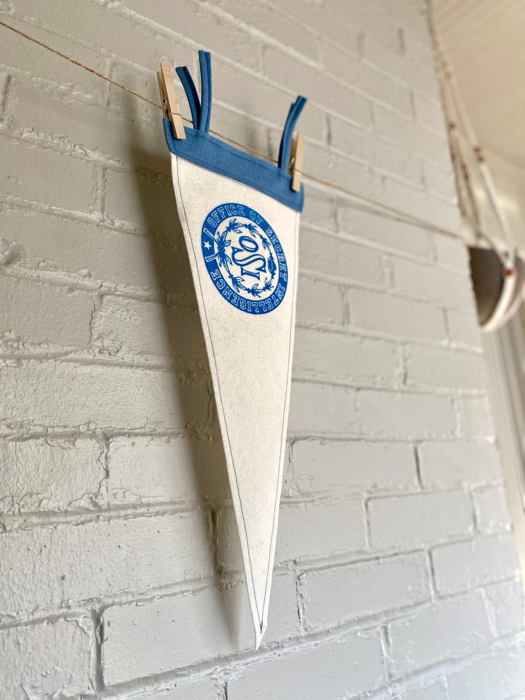 Side view of a white felt pennant with light blue trim and the OSI logo from the cartoon The Venture Bros printed in blue ink