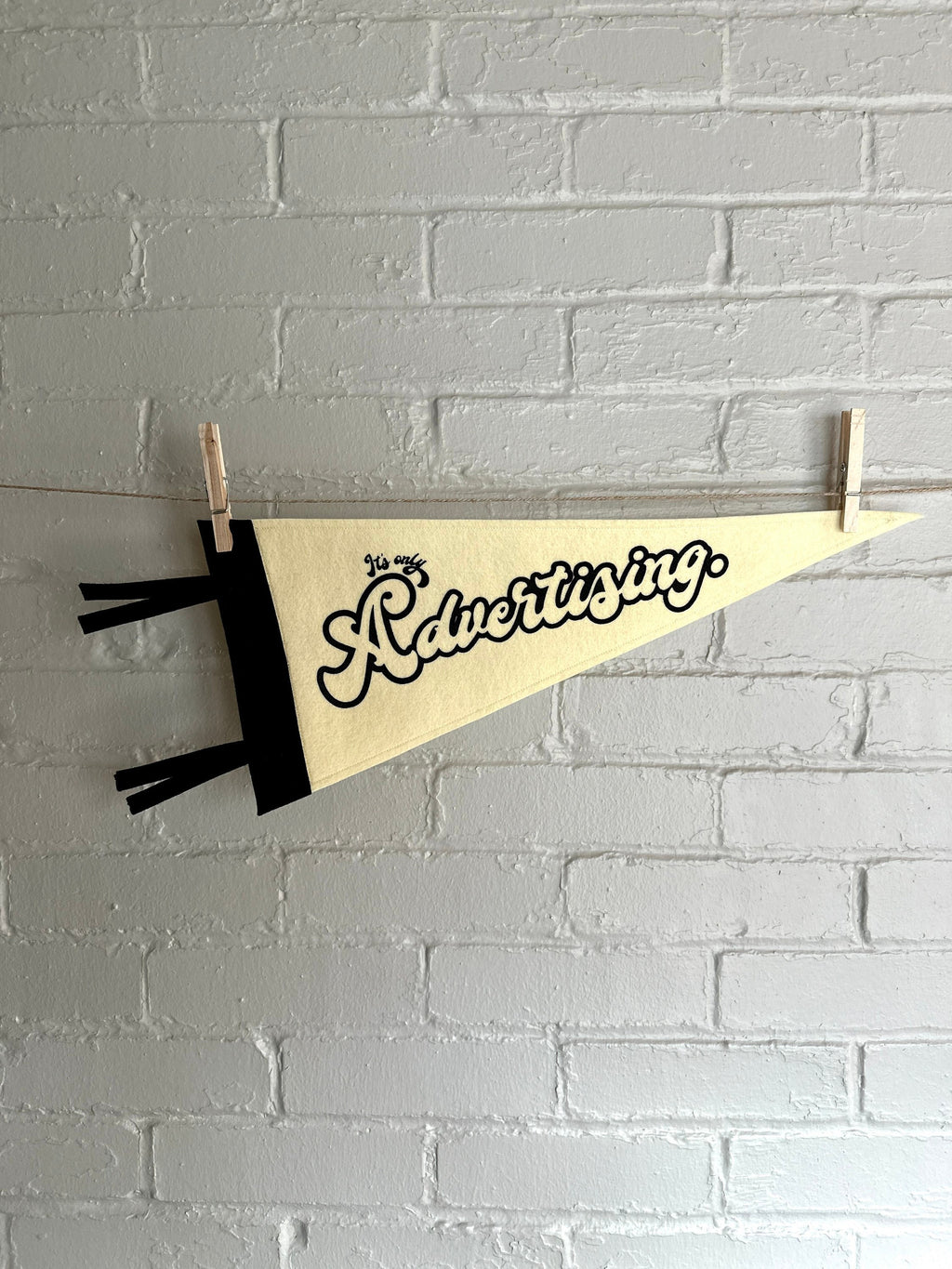 A cream felt pennant with black trim and print that reads "It's just advertising" hanging horizontally