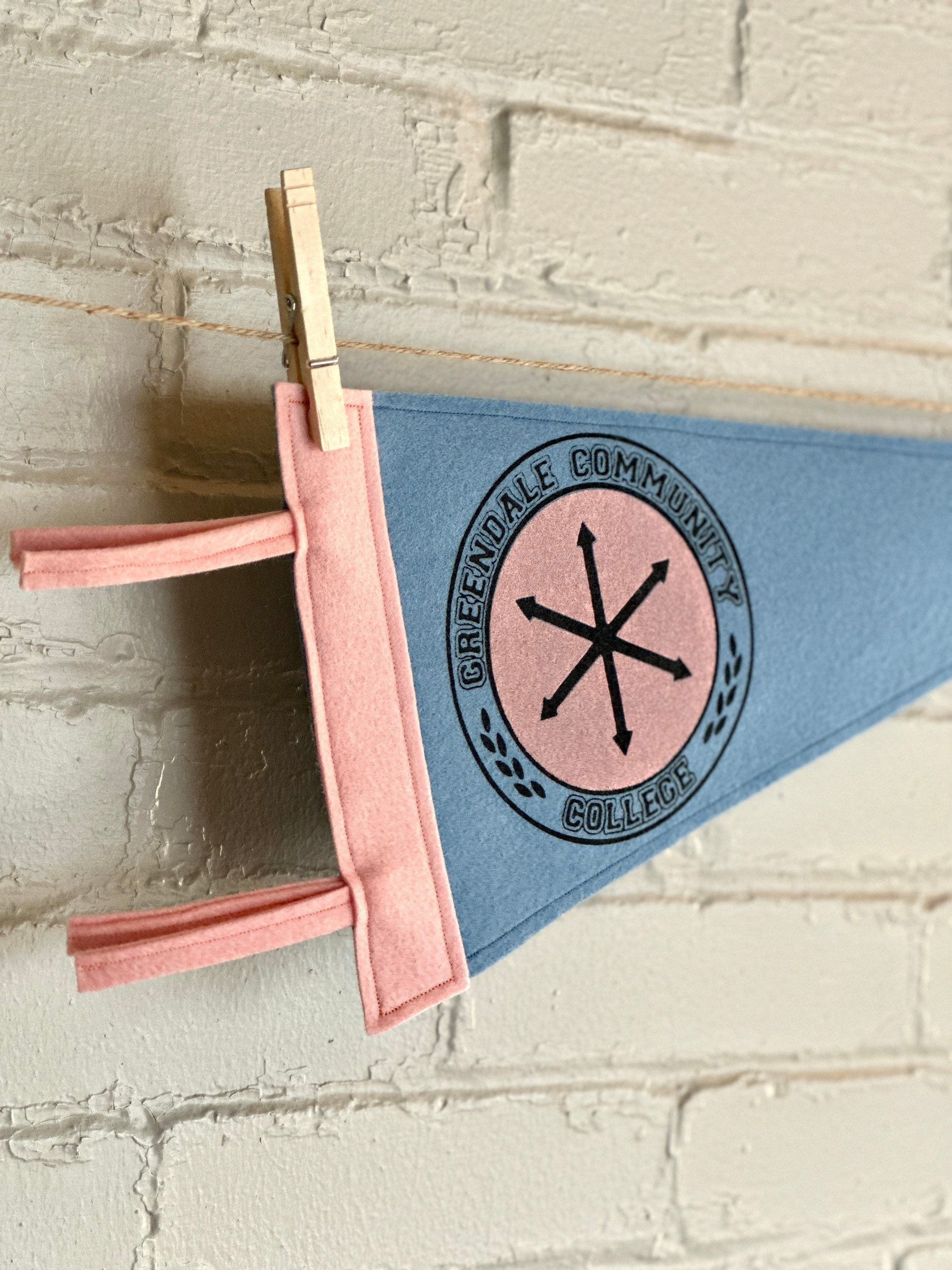 Closeup view of a light blue felt pennant with pink trim and a Greendale Community College logo printed on