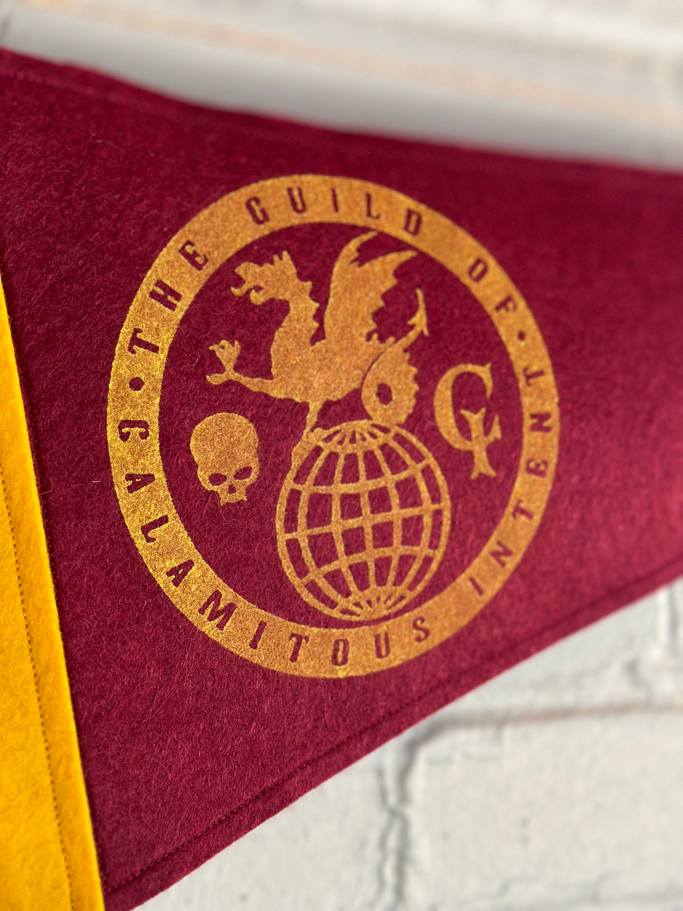 Close up side view of a burgundy pennant with yellow felt trim and the Guild of Calamitous Intent logo printed in yellow ink